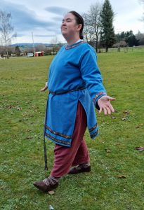 Ailith Ward wearing a mid blue tunic of cashmere, with decorative braid around the neck, wrists, with a narrow woven belt, over a pair of trousers, stripy socks and brown leather turn shoes