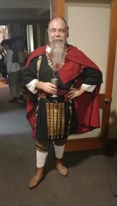 Angus wearing his black linen Roman tunic at Southron Gaard's SCA 60th Year Games Revel in AS 60.
Angus is wearing a black linen tunic with gold clavi over a natural linen undertunic, brown linen trousers, natural wool socks and brown leather boots. The ensemble is topped with a black edged cloak of red wool secured with a large brooch.