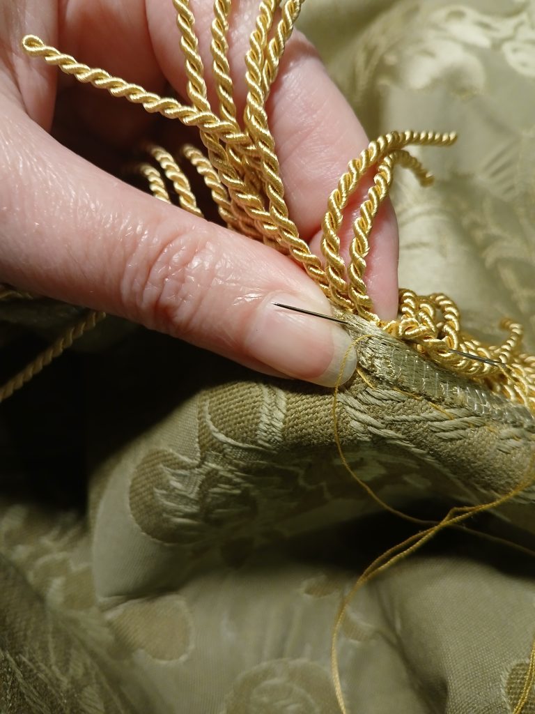 Gold fringe being hand stitched to the edge of a green brocade fabric